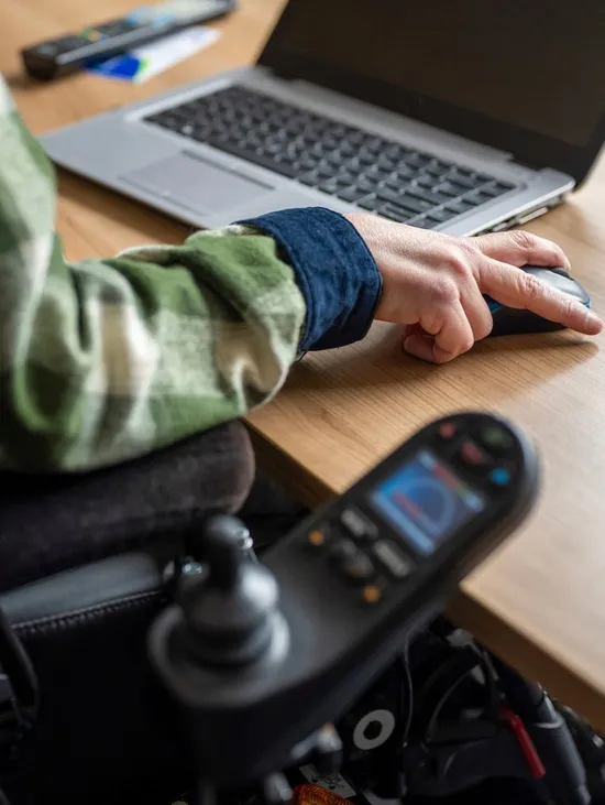 Nahaufnahme einer Person im Rollstuhl, die am Laptop arbeitet und eine Computermaus bedient.